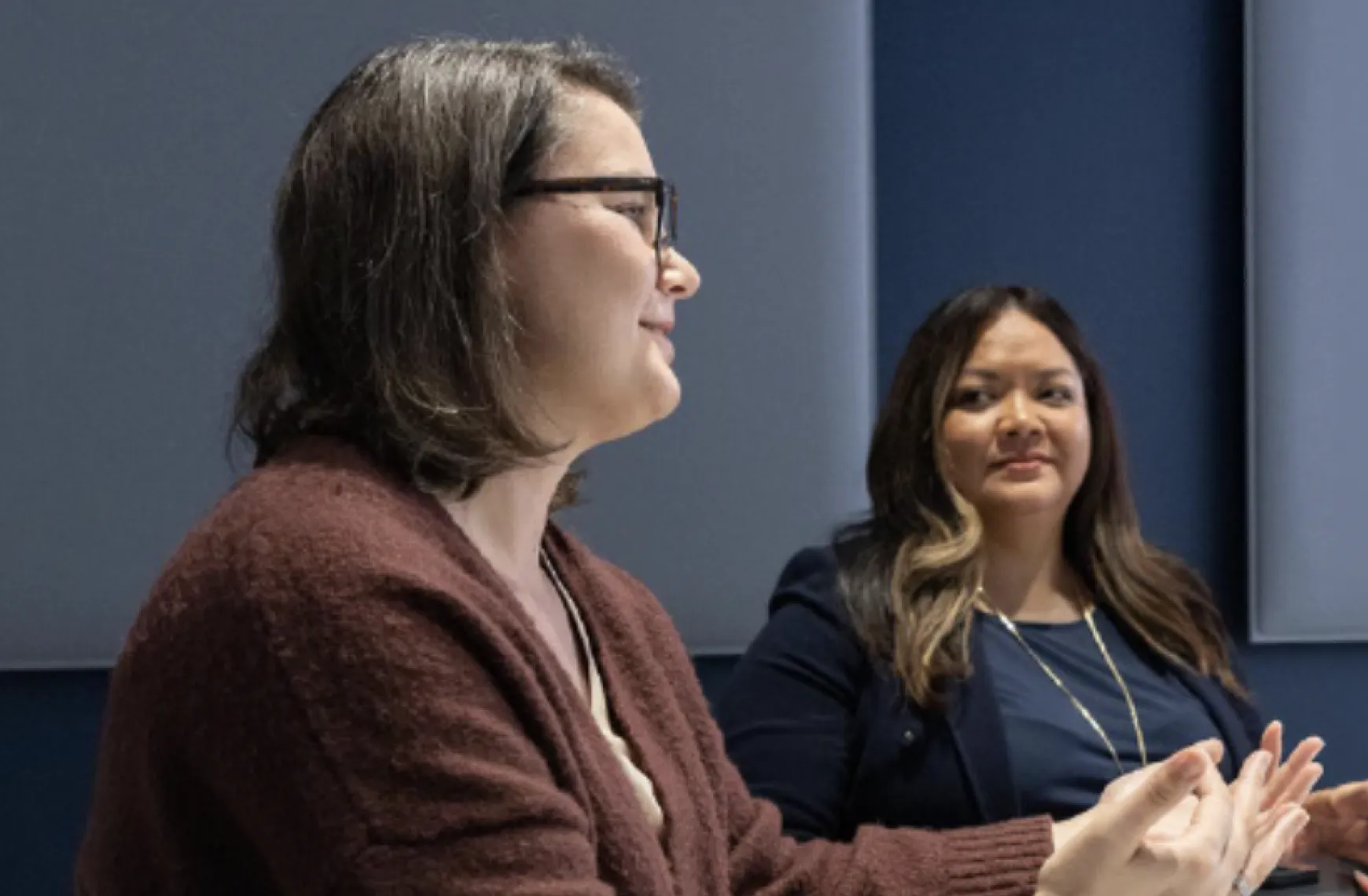 A woman wearing a brown sweater and glasses speaking while seated